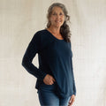 A woman with gray hair wearing a dark navy blue long-sleeve top and blue jeans, standing against a light beige textured wall.