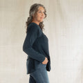 A woman with gray curly hair is shown in profile wearing a dark navy long-sleeve top and blue jeans, standing against a light beige textured fabric backdrop.