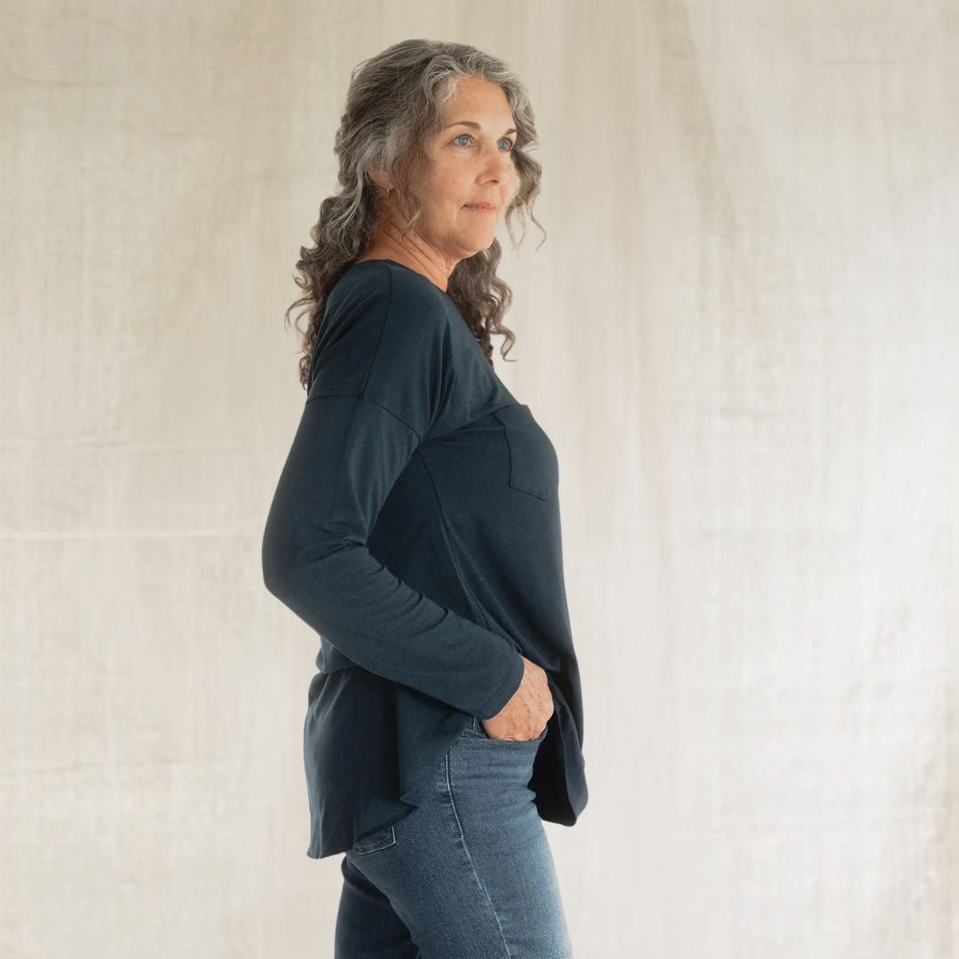 A woman with gray curly hair is shown in profile wearing a dark navy long-sleeve top and blue jeans, standing against a light beige textured fabric backdrop.