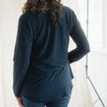A woman with long, wavy brown hair is shown from behind wearing a dark navy blue long-sleeve top and blue jeans, standing in front of a white curtain backdrop.
