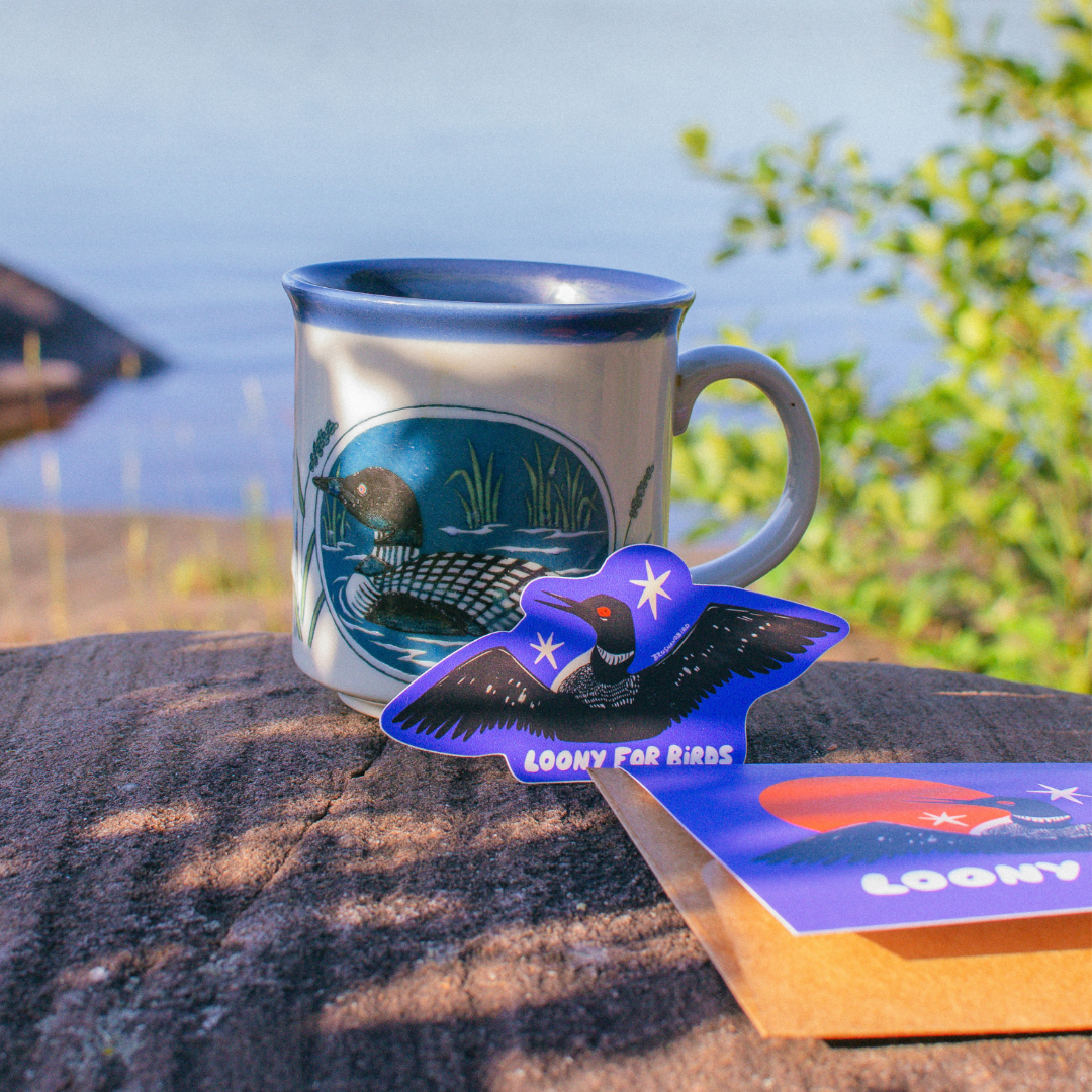 A ceramic mug with a blue rim and a loon illustration sits outdoors on a wooden surface, with a blue loon-shaped sticker and a small card in front of it. The background shows water, shoreline, and green foliage.