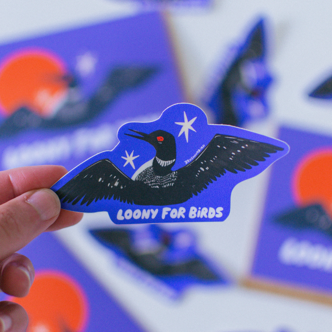 A hand holds a die-cut sticker featuring a black bird with white stars and the text 'LOONY FOR BIRDS' on a blue background, with more blurred stickers behind it.