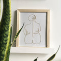 Framed abstract line art of a nude figure on a white wall with a plant in the foreground.