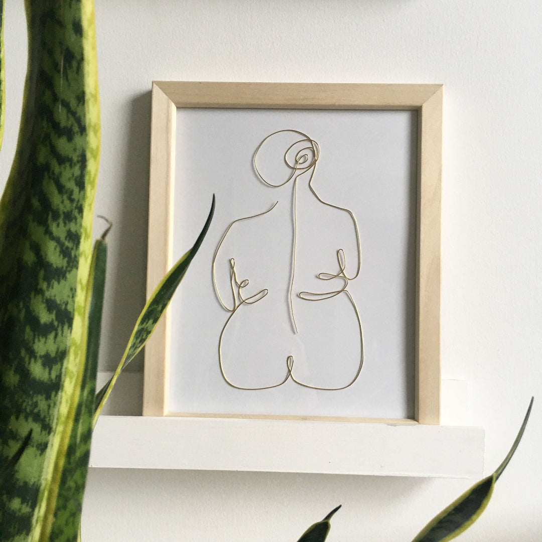 Framed abstract line art of a nude figure on a white wall with a plant in the foreground.