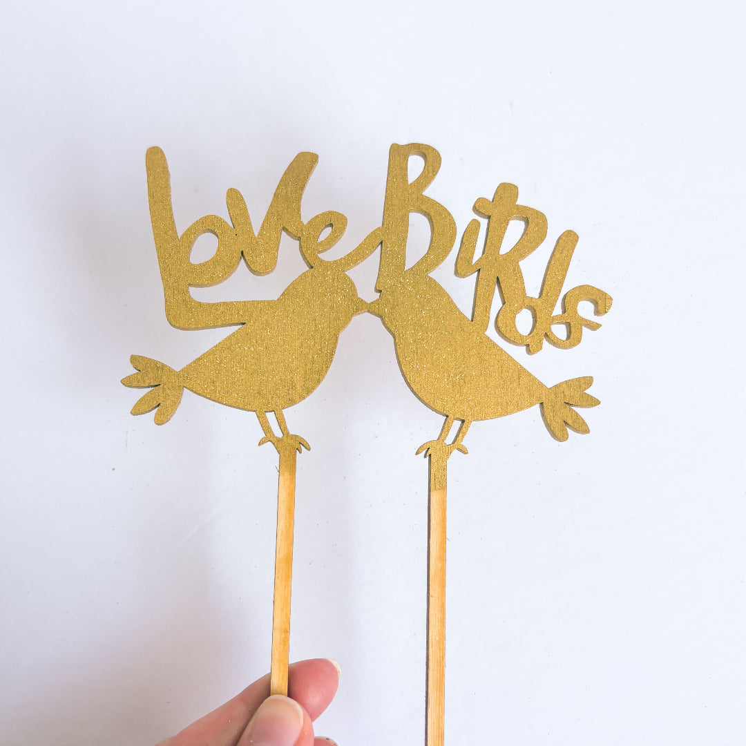 Gold 'Love Birds' cake toppers on sticks held by a hand against a white background