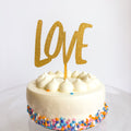 White cake with colorful sprinkles and a gold 'LOVE' topper on a white background