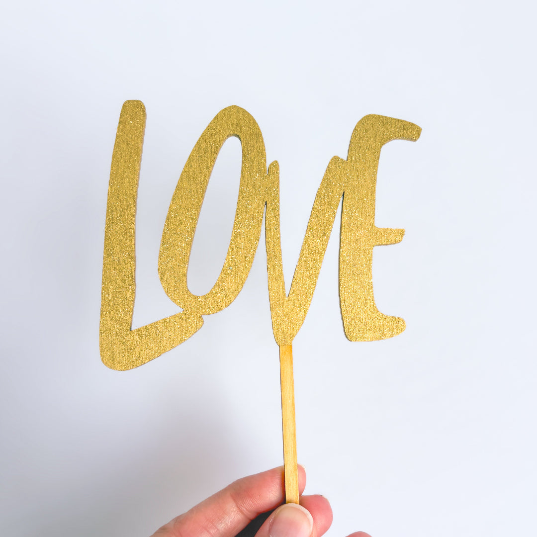 Gold glitter 'LOVE' cake topper held by a hand against a white background