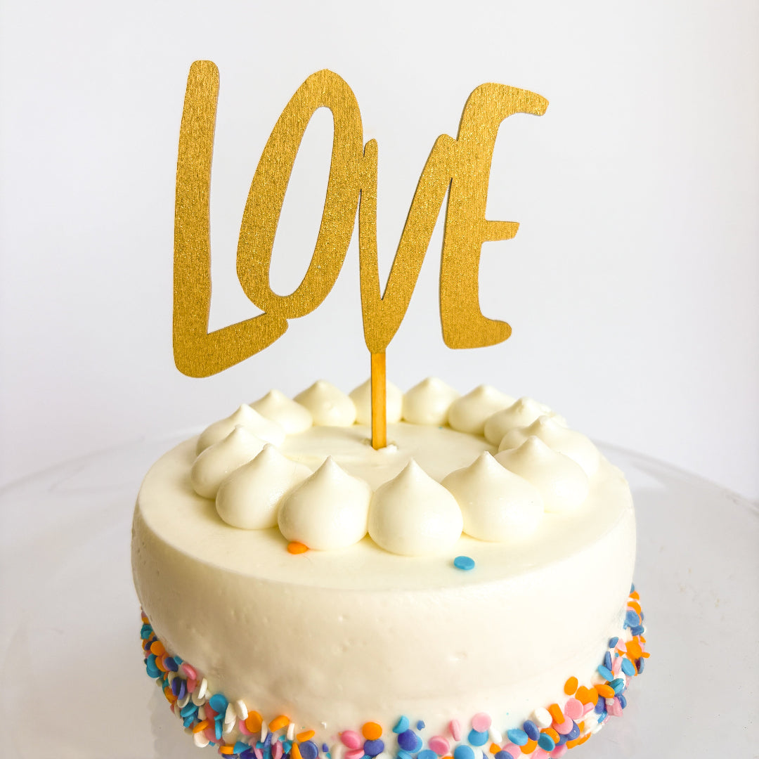 Small round cake with white frosting and colorful sprinkles, topped with a gold 'LOVE' cake topper on a white background.