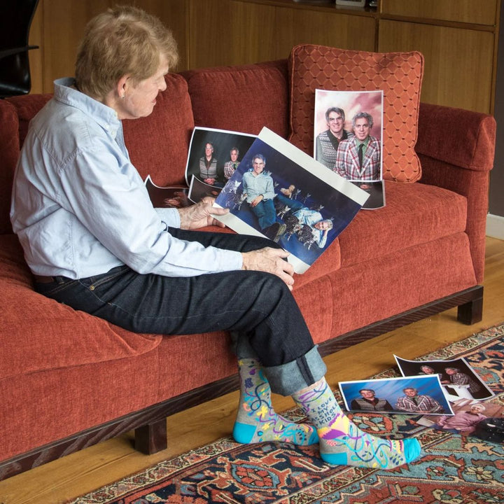 A pair of grey women's crew socks with vibrant, abstract floral and swirling line patterns in purple, yellow, pink, green, and blue. Text on one sock reads 