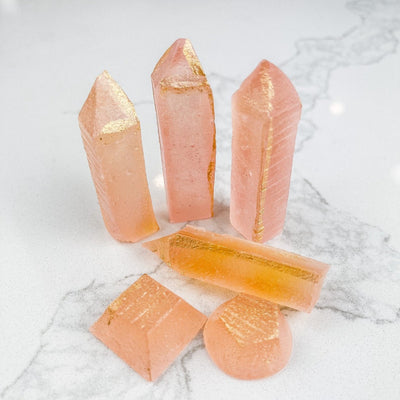 Several pink, crystal-shaped candies arranged on a white marble surface. Some candies have a pyramid shape, while others are more rounded. A few pieces have gold accents.
