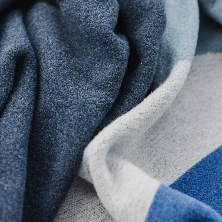 The River Reversible Blanket by Mini Tipi, featuring flowing waves of blue, grey, black, and white, held up by two hands. This reversible blanket is available in Ottawa, Ontario, Canada.