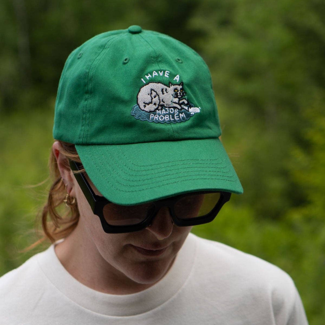 Person wearing a green cap with a design and sunglasses, standing in a forest.
