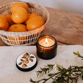 Candle with a coaster featuring an orange design, basket of oranges, and green leaves on a marble surface.