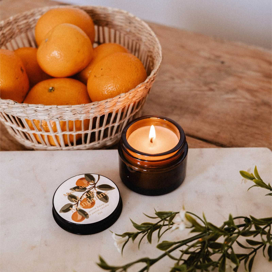Candle with a coaster featuring an orange design, basket of oranges, and green leaves on a marble surface.