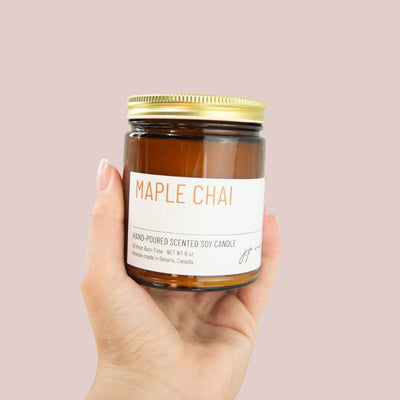 Hand holding a jar of 'Maple Chai' candle against a pink background
