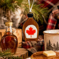 A maple syrup-shaped ornament with a red maple leaf on a white center hangs from a string in a cozy rustic setting with syrup bottles, pine branches, plaid fabric, and a mug.