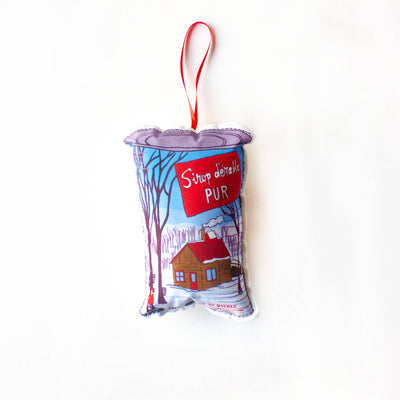 A close-up, high-angle flat lay shot on a plain white background features a soft, plush Christmas ornament shaped like a can of maple syrup. The can has a blue and red label with a cartoon winter scene showing a small wooden cabin among trees. A red oval banner in the center reads 