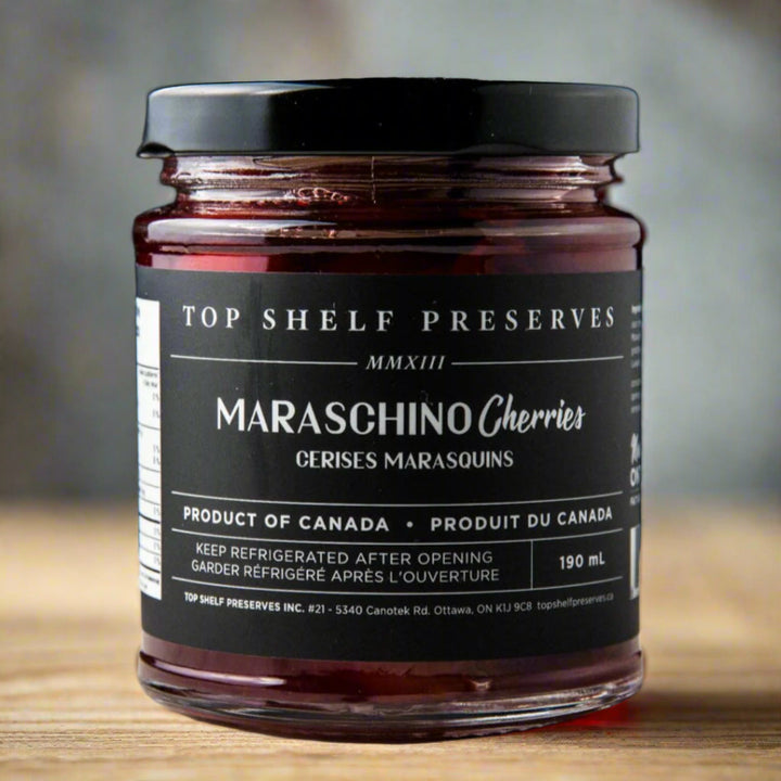 A jar of Maraschino Cherries from Top Shelf Preserves. The jar is filled with red cherries and has a black label with white text. The label includes the product name, 