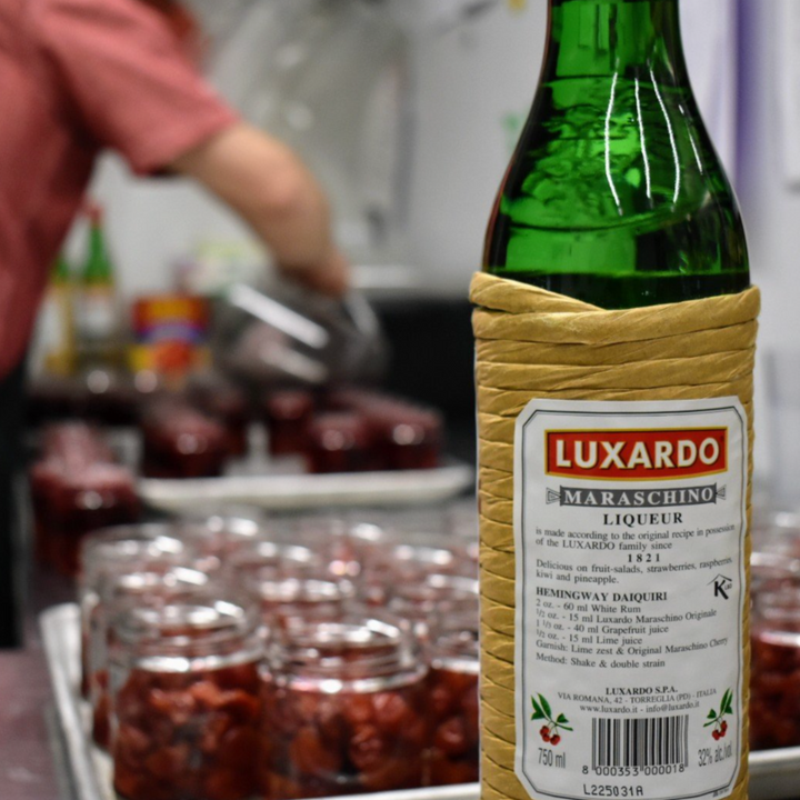A jar of Maraschino Cherries from Top Shelf Preserves. The jar is filled with red cherries and has a black label with white text. The label includes the product name, 