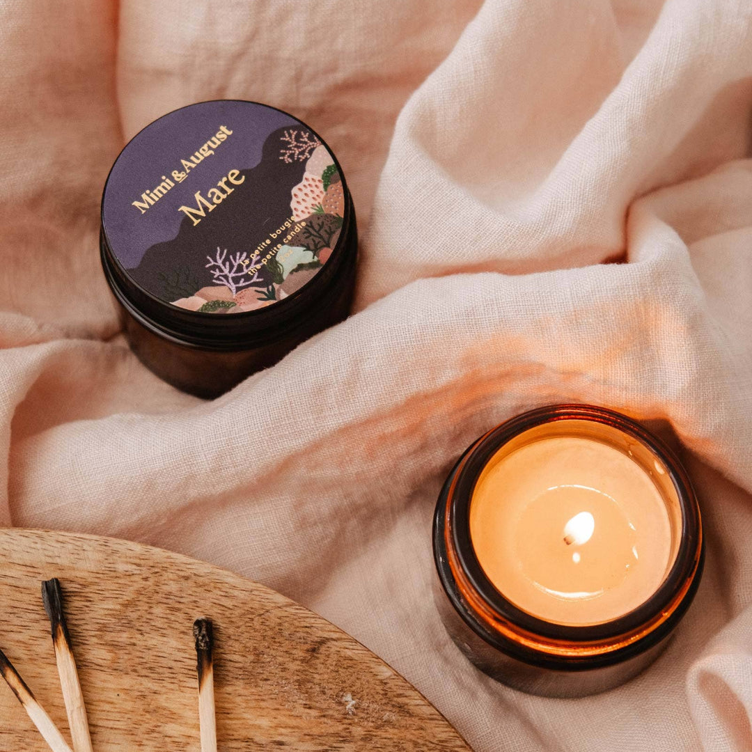 Candle with a lit wick on a soft fabric background, next to a container labeled 'Mare'.