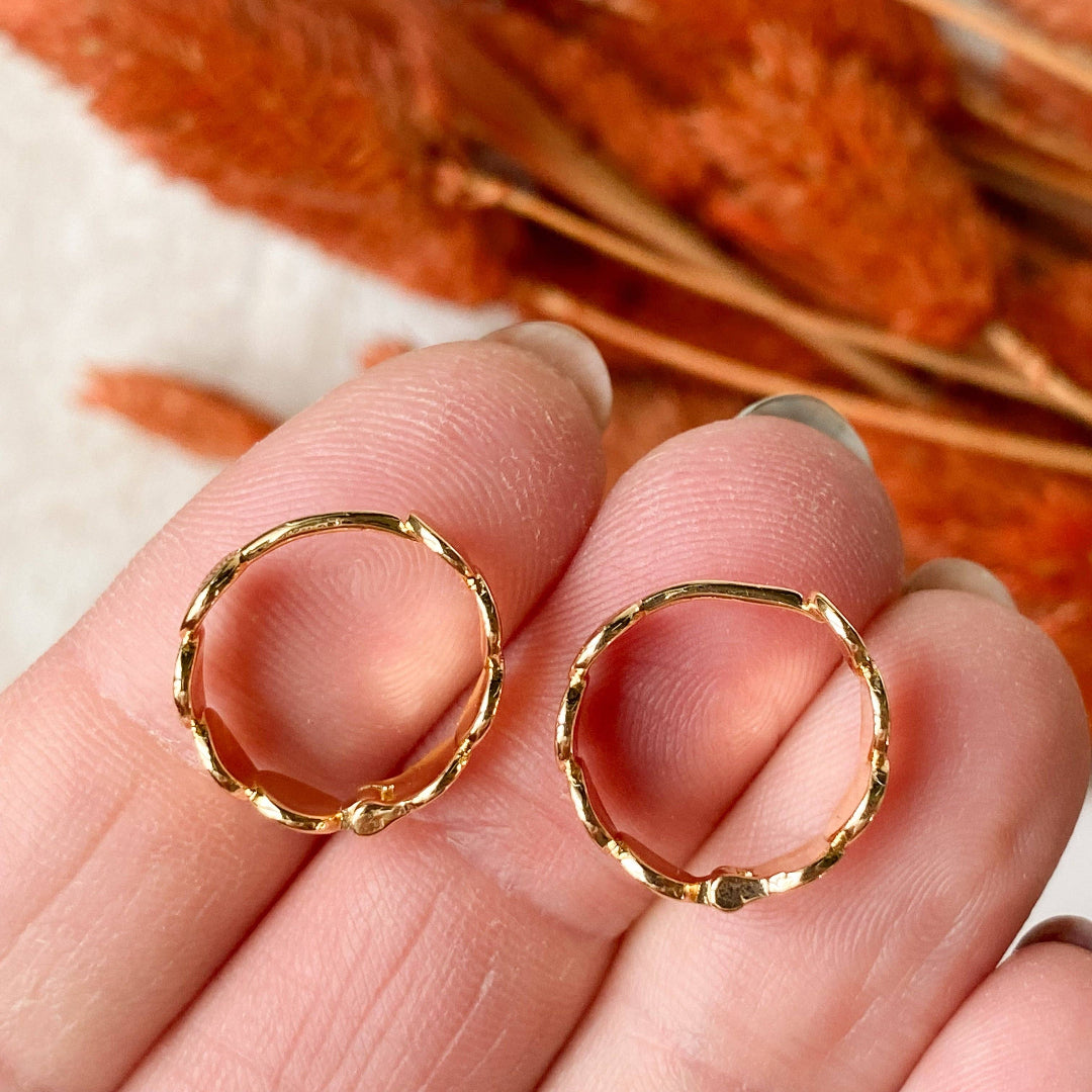 Gold hoop earrings held between fingers with a blurred background of orange flowers.