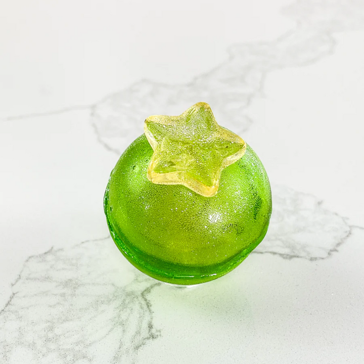 A green, spherical tea bomb topped with a yellow star. The tea bomb is resting on a white marble surface.