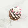 A hot chocolate bomb decorated with white chocolate drizzle, a white butterfly, and pink sugar beads. The bomb is resting on a white marble surface.