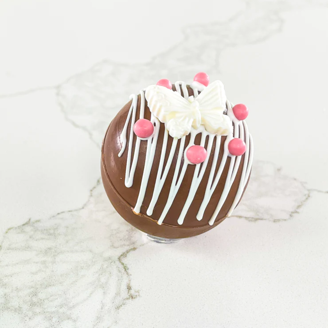 A hot chocolate bomb decorated with white chocolate drizzle, a white butterfly, and pink sugar beads. The bomb is resting on a white marble surface.