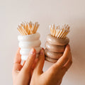 Two hands holding small ceramic containers with toothpicks against a plain background