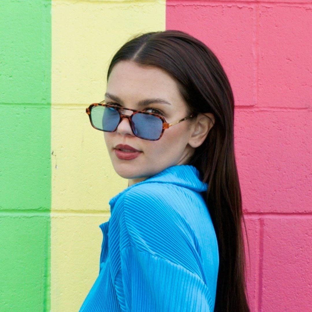 Woman wearing sunglasses and a blue top against a colorful striped wall.