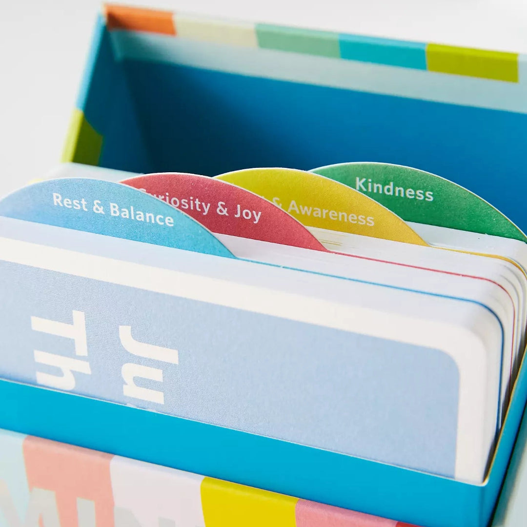 A close-up of a colorful card deck in a matching box, showing card tabs labeled 'Rest & Balance,' 'compassion & Joy,' 'Awareness,' and 'Kindness.'.