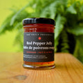 A jar of Red Pepper Jelly from Top Shelf Preserves. The jar is filled with a red jelly and has a black label with white text. The label includes the product name, "Handmade Fait à la Main," and instructions to keep refrigerated after opening.