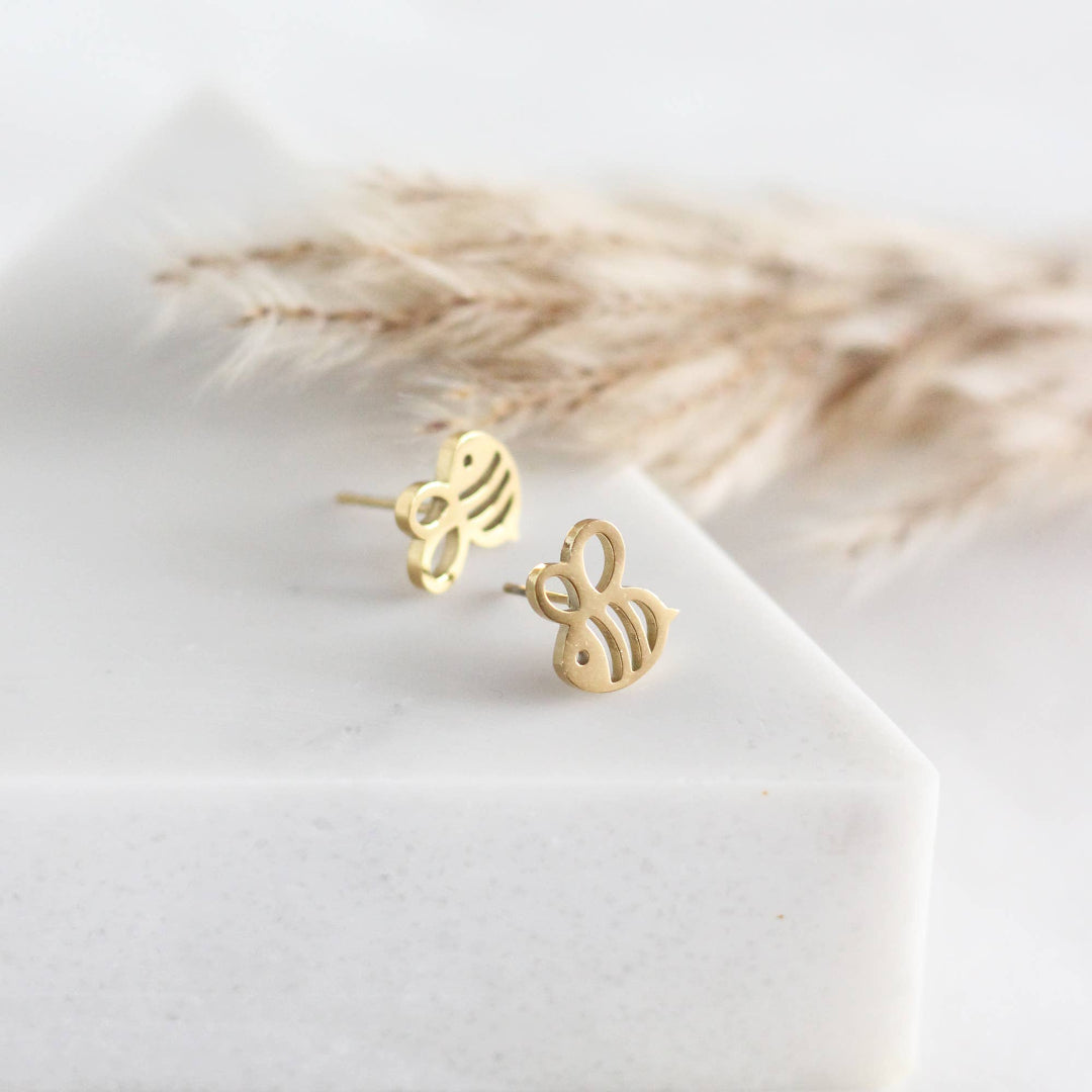 Gold bee-shaped earrings on a white surface with dried plants in the background