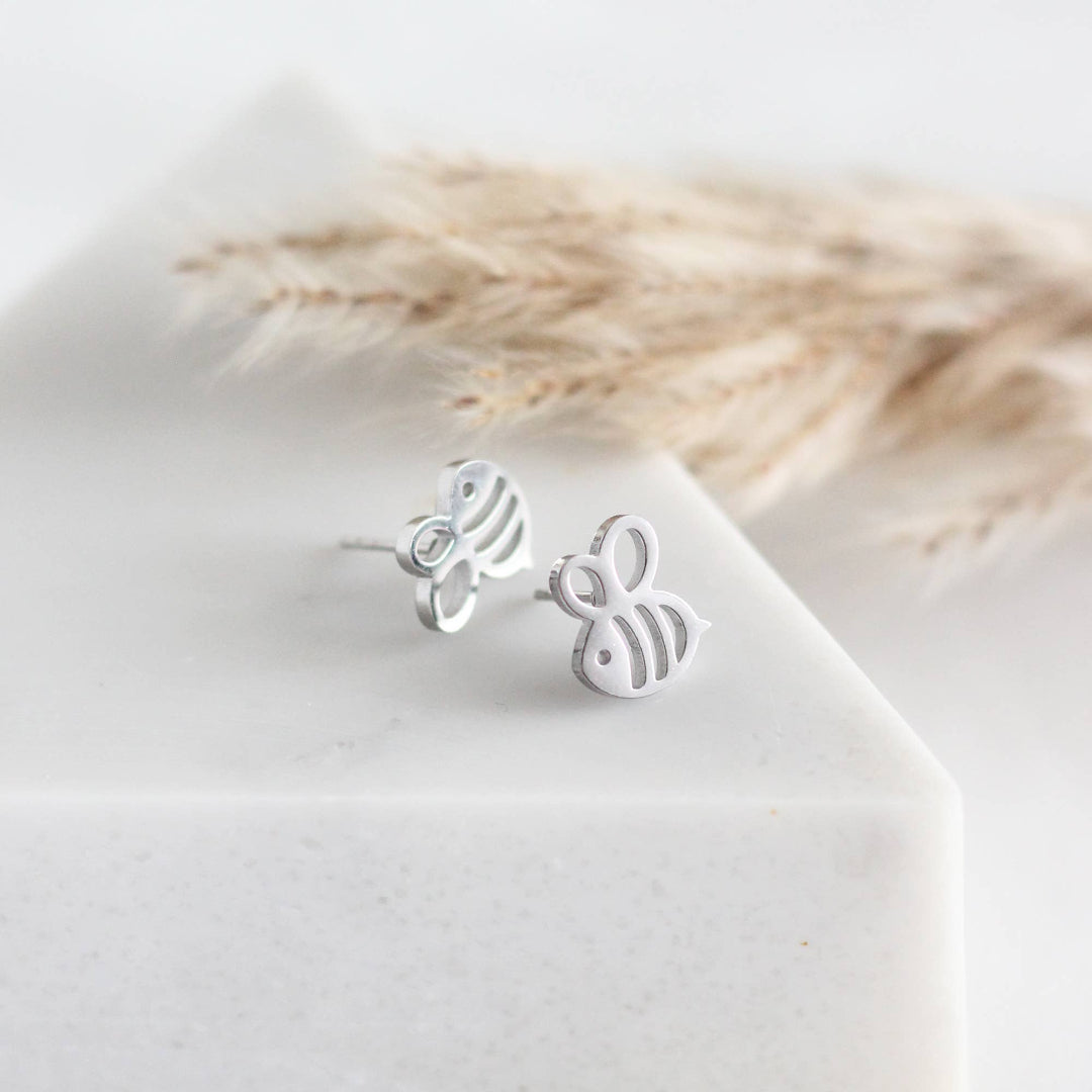 Silver bee-shaped earrings on a white surface with dried plants in the background