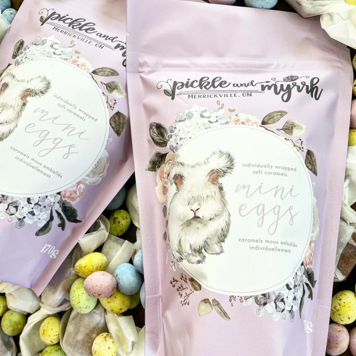 A purple bag of Pickle + Myrrh Mini Egg Caramels with a drawing of a bunny and the text 'mini eggs', surrounded by loose mini egg caramels in various pastel colors. These are gourmet, handmade caramels from Merrickville, Ontario, Canada.