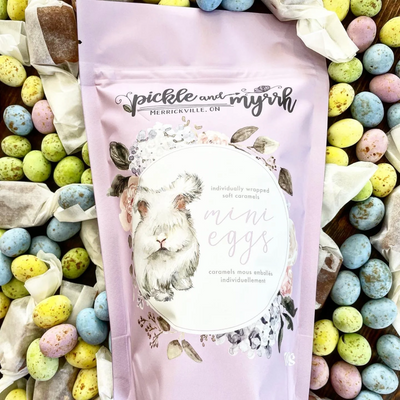A purple bag of Pickle + Myrrh Mini Egg Caramels with a drawing of a bunny and the text 'mini eggs', surrounded by loose mini egg caramels in various pastel colors. These are gourmet, handmade caramels from Merrickville, Ontario, Canada.