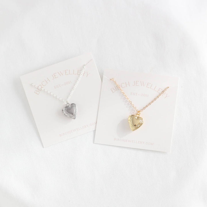 Two heart-shaped locket necklaces on a white card. One necklace has a gold-colored locket, and the other has a silver-colored locket. Both lockets have detailed designs and are attached to delicate chains.