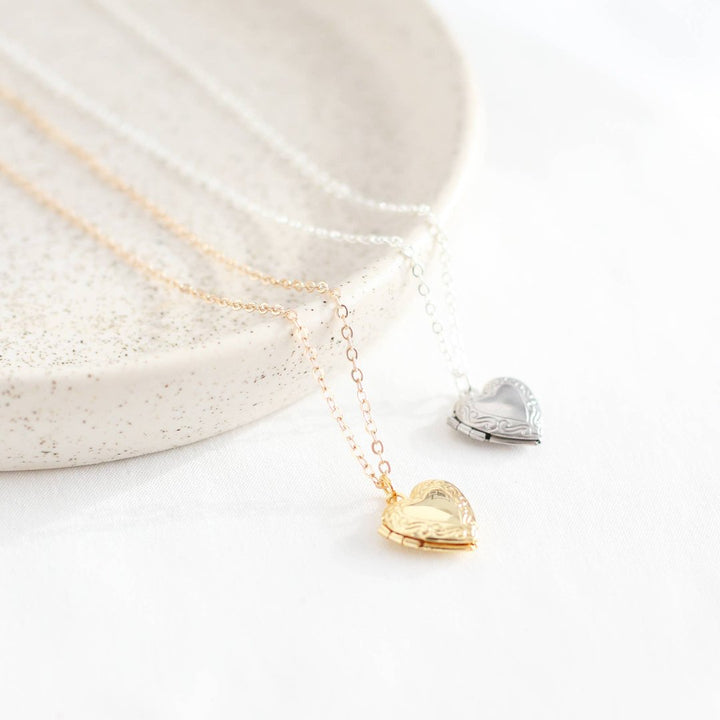 Two heart-shaped locket necklaces on a white card. One necklace has a gold-colored locket, and the other has a silver-colored locket. Both lockets have detailed designs and are attached to delicate chains.