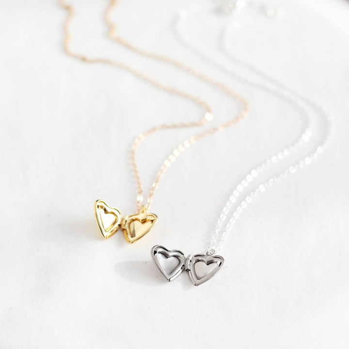 Two heart-shaped locket necklaces on a white card. One necklace has a gold-colored locket, and the other has a silver-colored locket. Both lockets have detailed designs and are attached to delicate chains.