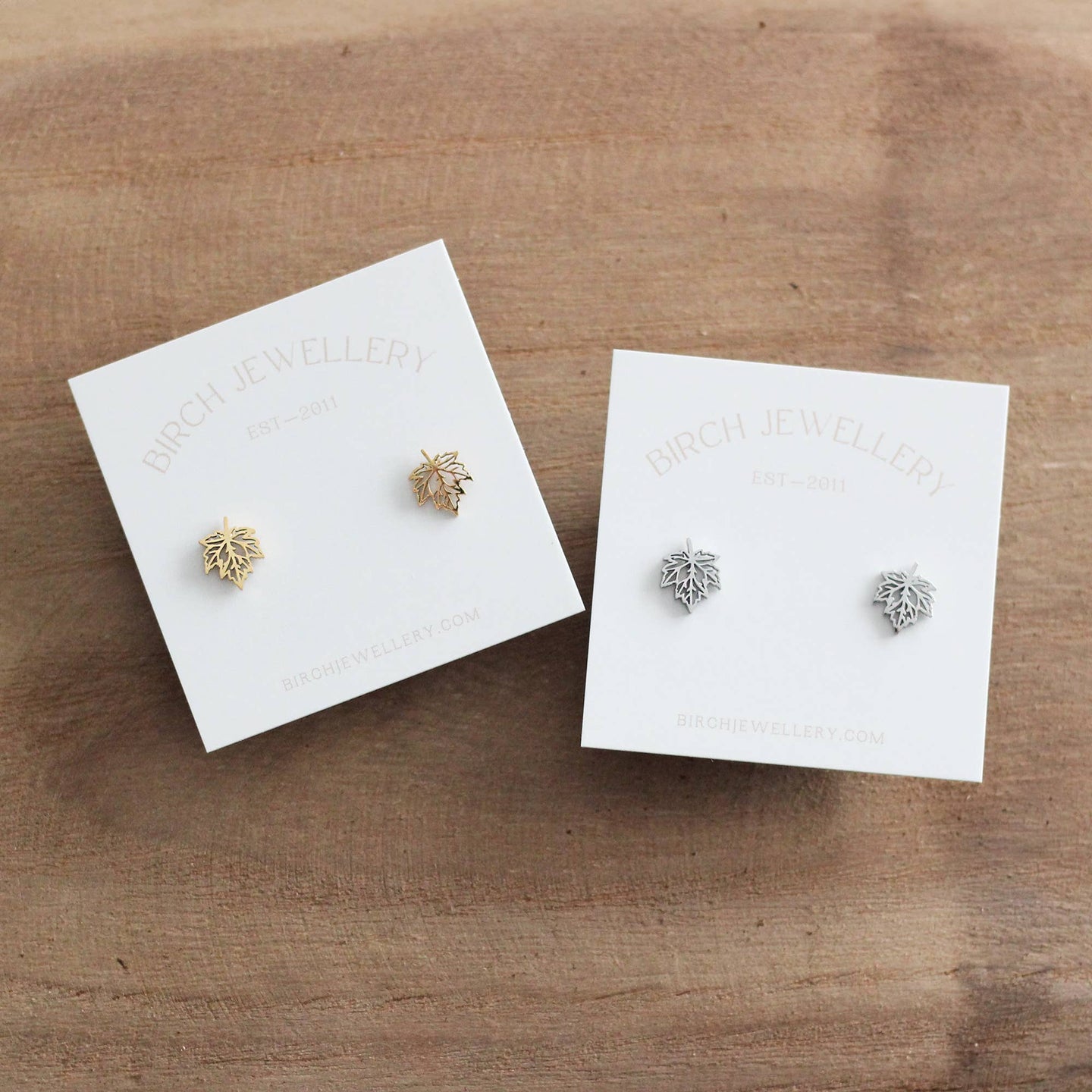 Two pairs of maple leaf shaped earrings on white cards with 'Birch & Willow' branding on a wooden surface.