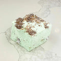 A square marshmallow with swirls of mint green and brown chocolate chips. The marshmallow is resting on a white marble surface.