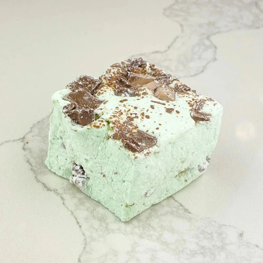 A square marshmallow with swirls of mint green and brown chocolate chips. The marshmallow is resting on a white marble surface.