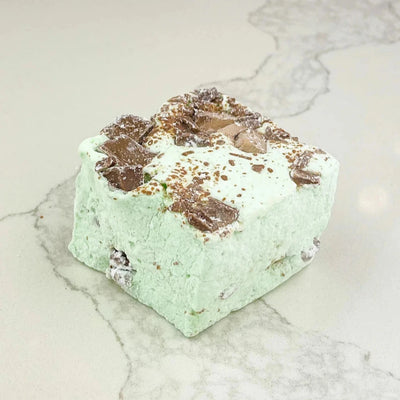 A square marshmallow with swirls of mint green and brown chocolate chips. The marshmallow is resting on a white marble surface.