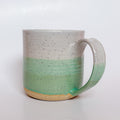 A handmade ceramic mug with a colorblock design featuring a white top and a speckled green middle section, displayed on a light gray background. The mug has a large, rounded handle, a speckled texture, and is made in Mississippi Mills, Ontario, Canada.