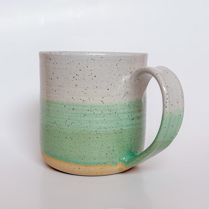 A handmade ceramic mug with a colorblock design featuring a white top and a speckled green middle section, displayed on a light gray background. The mug has a large, rounded handle, a speckled texture, and is made in Mississippi Mills, Ontario, Canada.