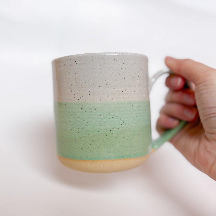 A handmade ceramic mug with a colorblock design featuring a white top and a speckled green middle section, displayed on a light gray background. The mug has a large, rounded handle, a speckled texture, and is made in Mississippi Mills, Ontario, Canada.