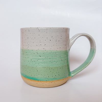 A handmade ceramic mug with a colorblock design featuring a white top and a speckled green middle section, displayed on a light gray background. The mug has a large, rounded handle, a speckled texture, and is made in Mississippi Mills, Ontario, Canada.
