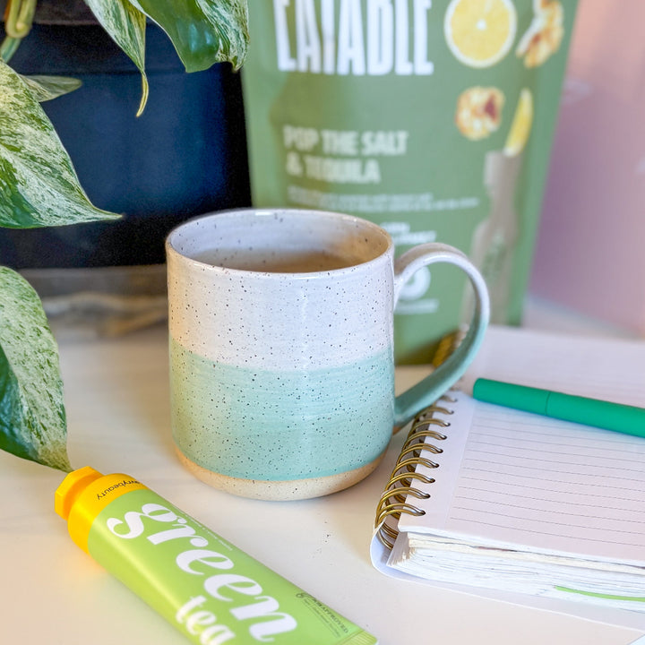 A handmade ceramic mug with a colorblock design featuring a white top and a speckled green middle section, displayed on a light gray background. The mug has a large, rounded handle, a speckled texture, and is made in Mississippi Mills, Ontario, Canada.