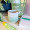 A handmade ceramic mug with a colorblock design featuring a white top and a speckled green middle section, displayed on a light gray background. The mug has a large, rounded handle, a speckled texture, and is made in Mississippi Mills, Ontario, Canada.