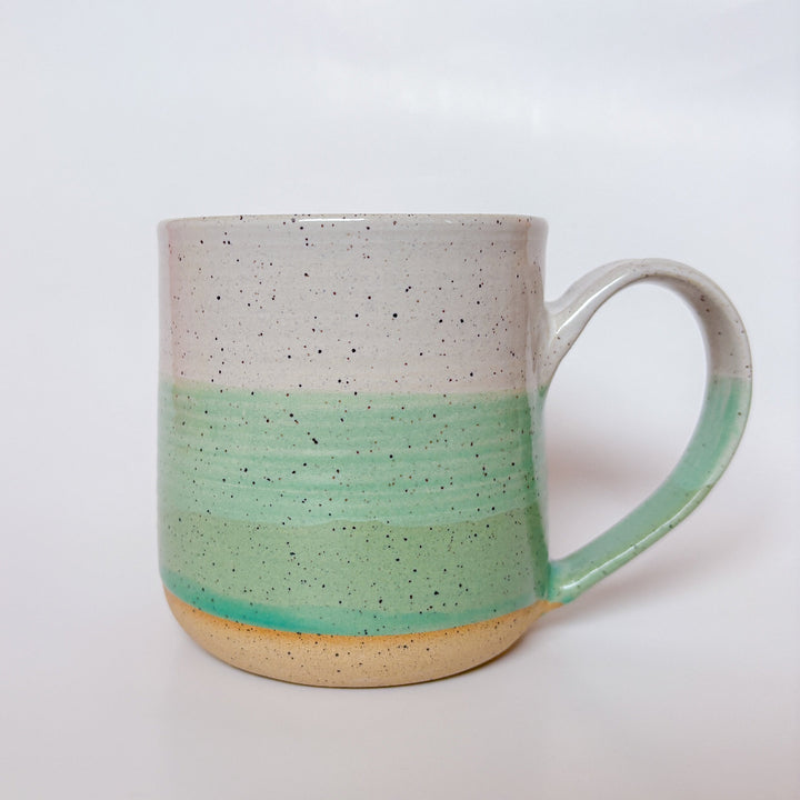 A handmade ceramic mug with a colorblock design featuring a white top and a speckled green middle section, displayed on a light gray background. The mug has a large, rounded handle, a speckled texture, and is made in Mississippi Mills, Ontario, Canada.